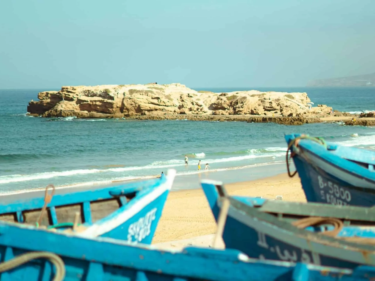 Praia em Marrocos com barcos azuis e rochas ao fundo.