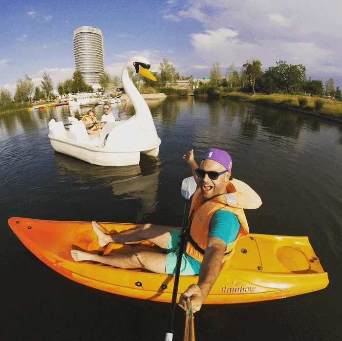 Hombre en kayak naranja sonríe mientras navega junto a un cisne en un lago.