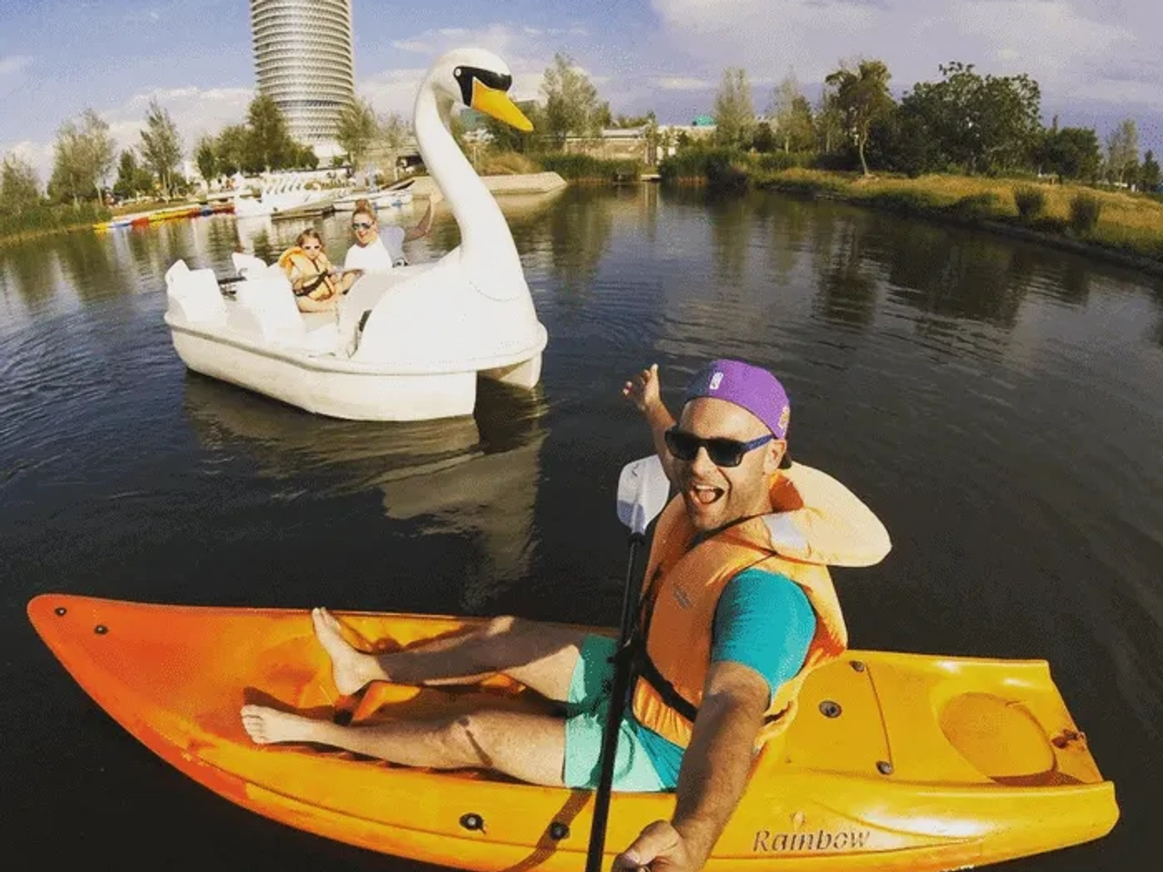 Hombre en kayak naranja sonríe mientras navega junto a un cisne en un lago.