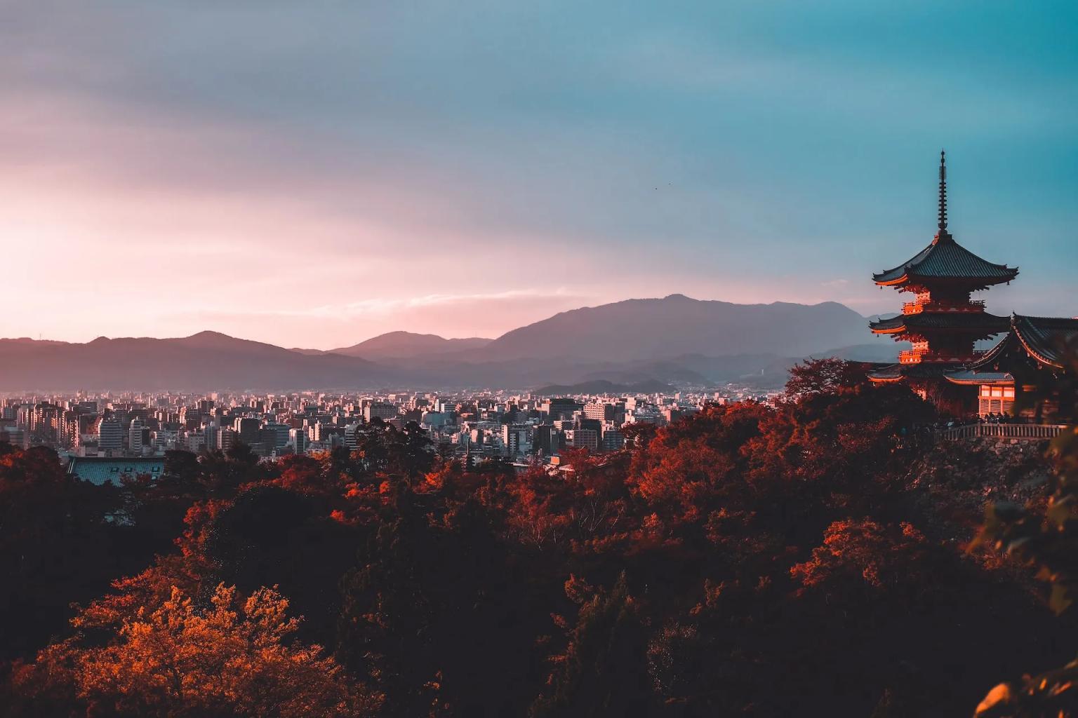 A traditional pagoda overlooks a cityscape with mountains in the background at sunset.