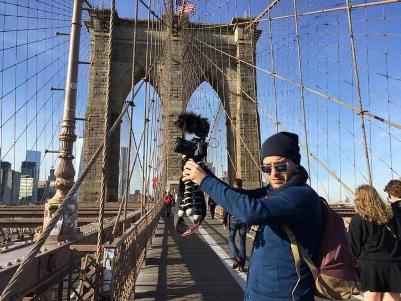 Hombre con cámara en el Puente de Brooklyn, fondo urbano y cielo despejado.