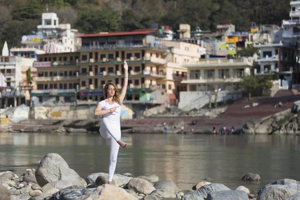Mujer practicando yoga en una playa con edificios al fondo.