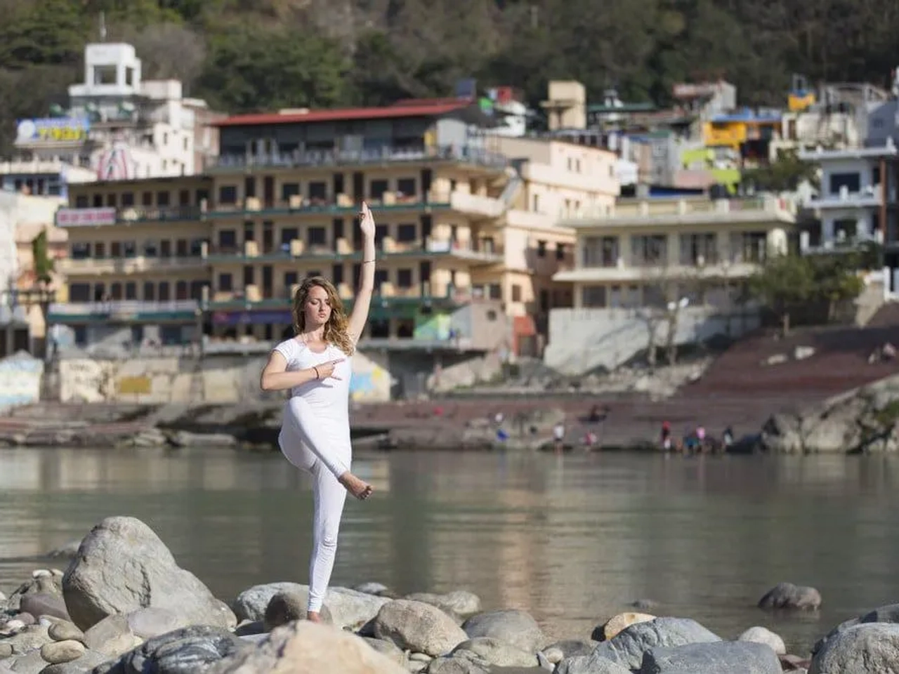 Mujer practicando yoga en una playa con edificios al fondo.