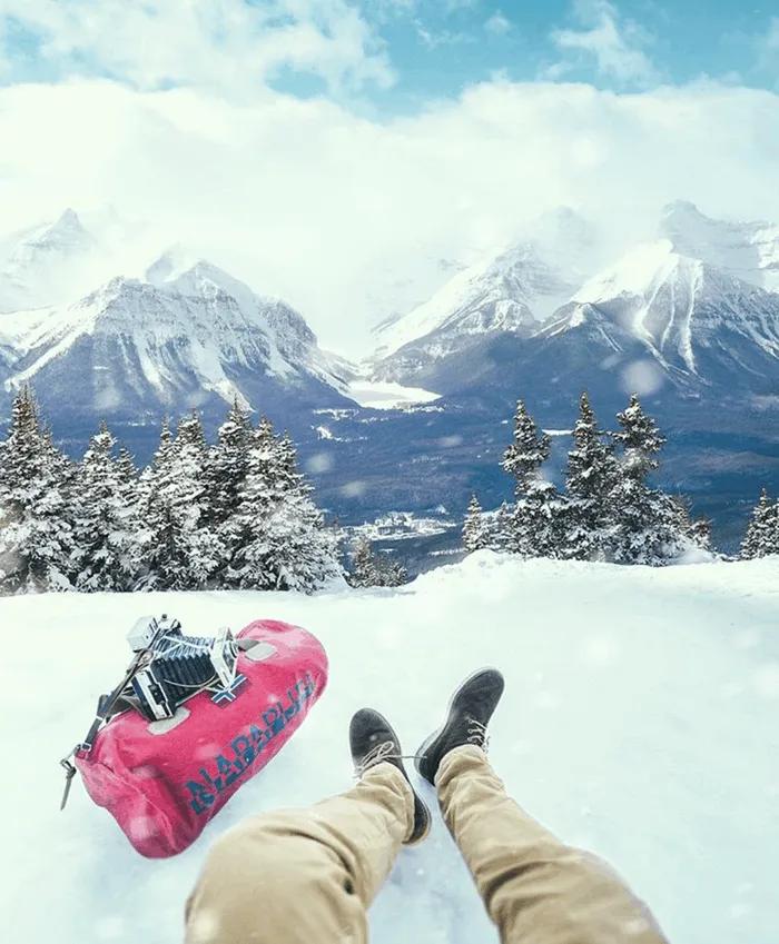 Persona sentada en la nieve, con montañas nevadas y un bolso de viaje rosa.
