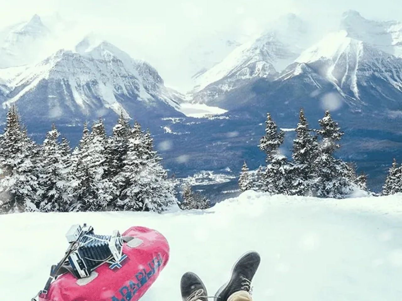 Persona sentada en la nieve, con montañas nevadas y un bolso de viaje rosa.