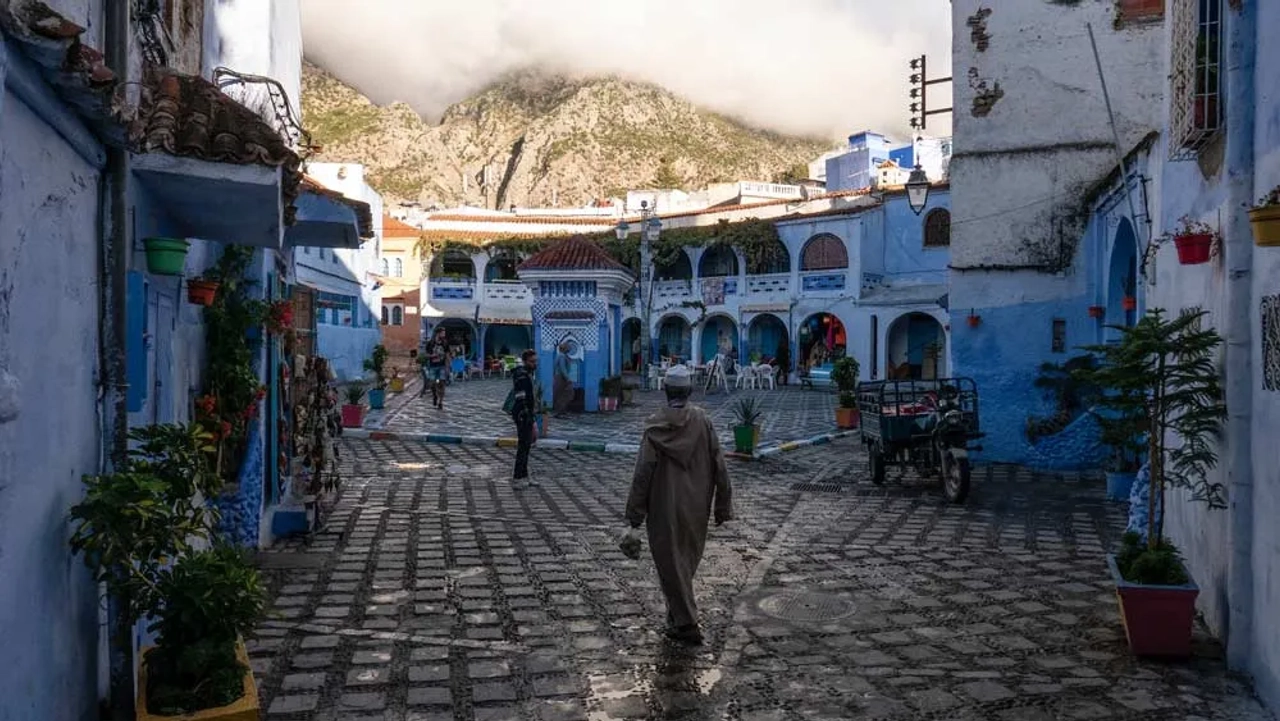 Piazza nella medina blu di Chefchaouen, con edifici bianchi e azzurri, montagne sullo sfondo e una persona che cammina tra le strade acciottolate.