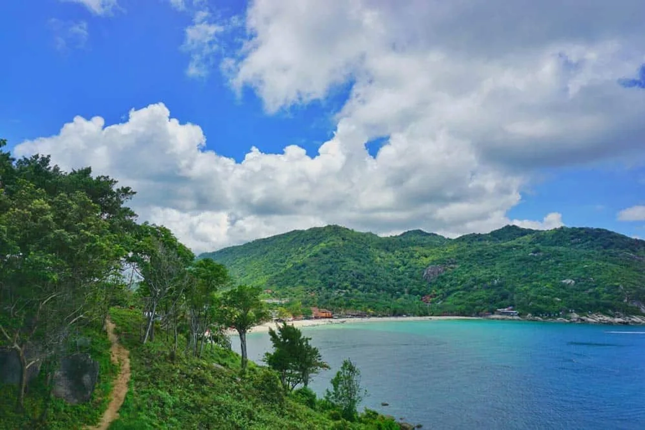 Vista panoramica della costa di Koh Phangan, con mare azzurro, spiaggia, colline verdi e un sentiero immerso nella vegetazione tropicale.