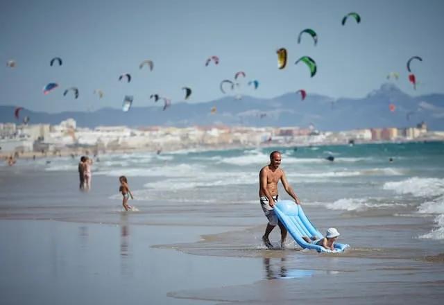 pessoas na praia, com muitos kitesurfs no mar e um pai a brincar com uma criança nas ondas