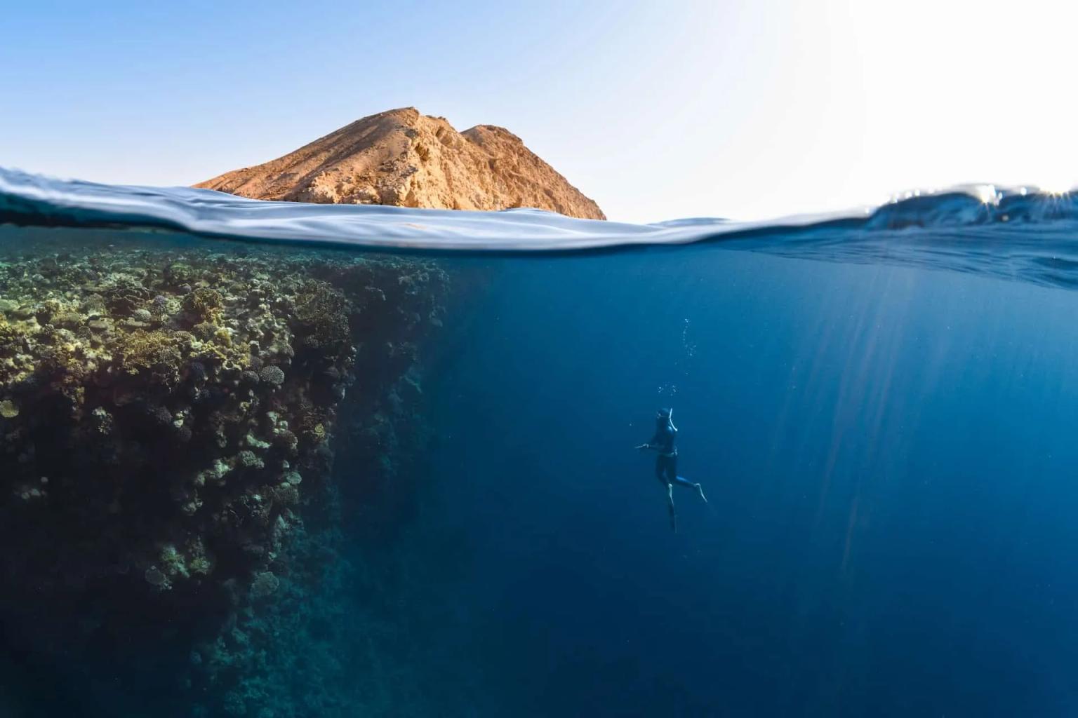 Mergulhador explorando recifes de corais em águas cristalinas, com montanha ao fundo.