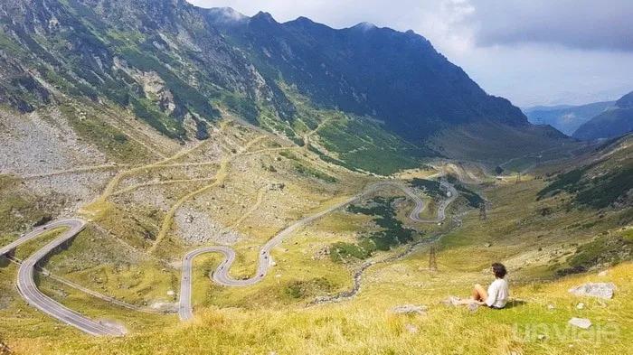 Vista panorámica de montañas y carretera serpenteante, con una persona sentada en el paisaje.