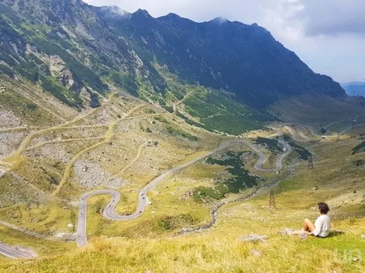 Vista panorámica de montañas y carretera serpenteante, con una persona sentada en el paisaje.