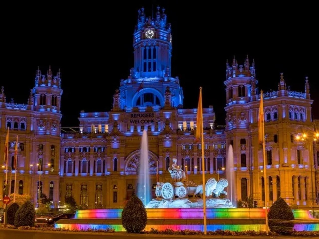 Edificio iluminado en colores, con fuente y mensaje de bienvenida a refugiados.