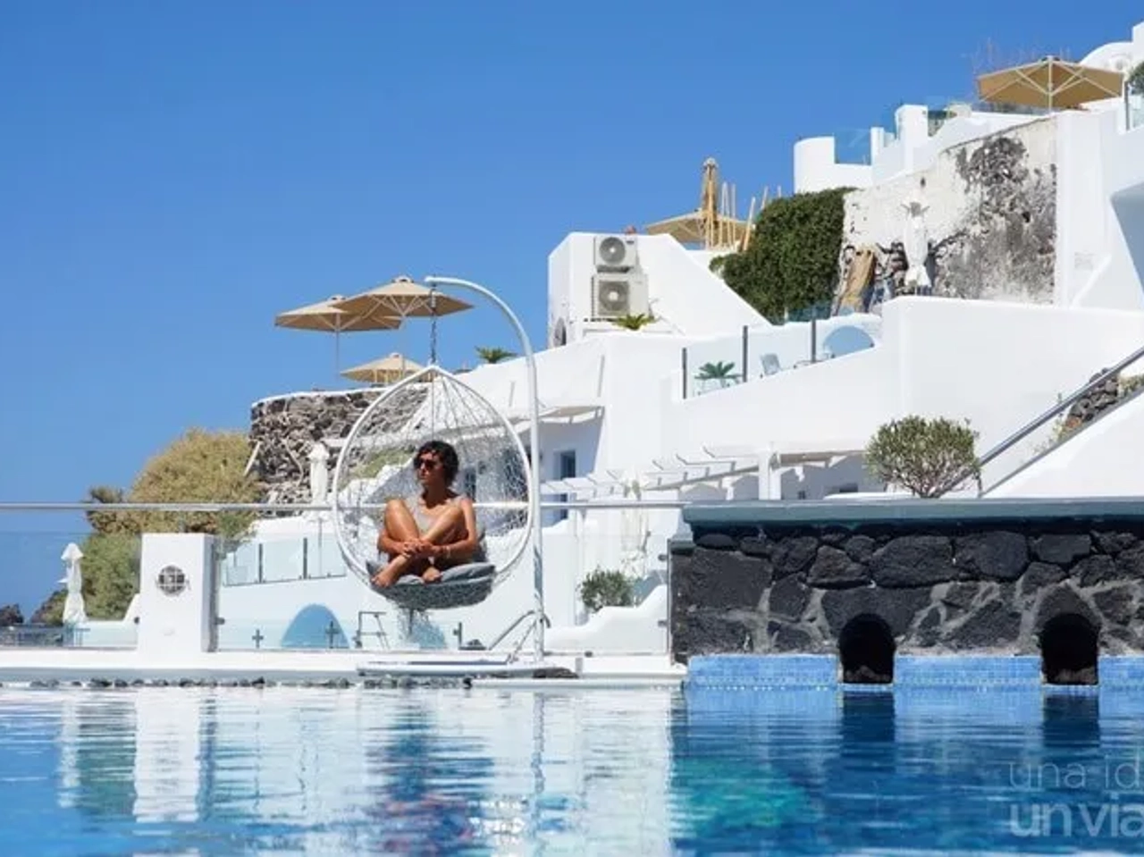 Persona sentada en una silla colgante junto a una piscina en un entorno soleado.