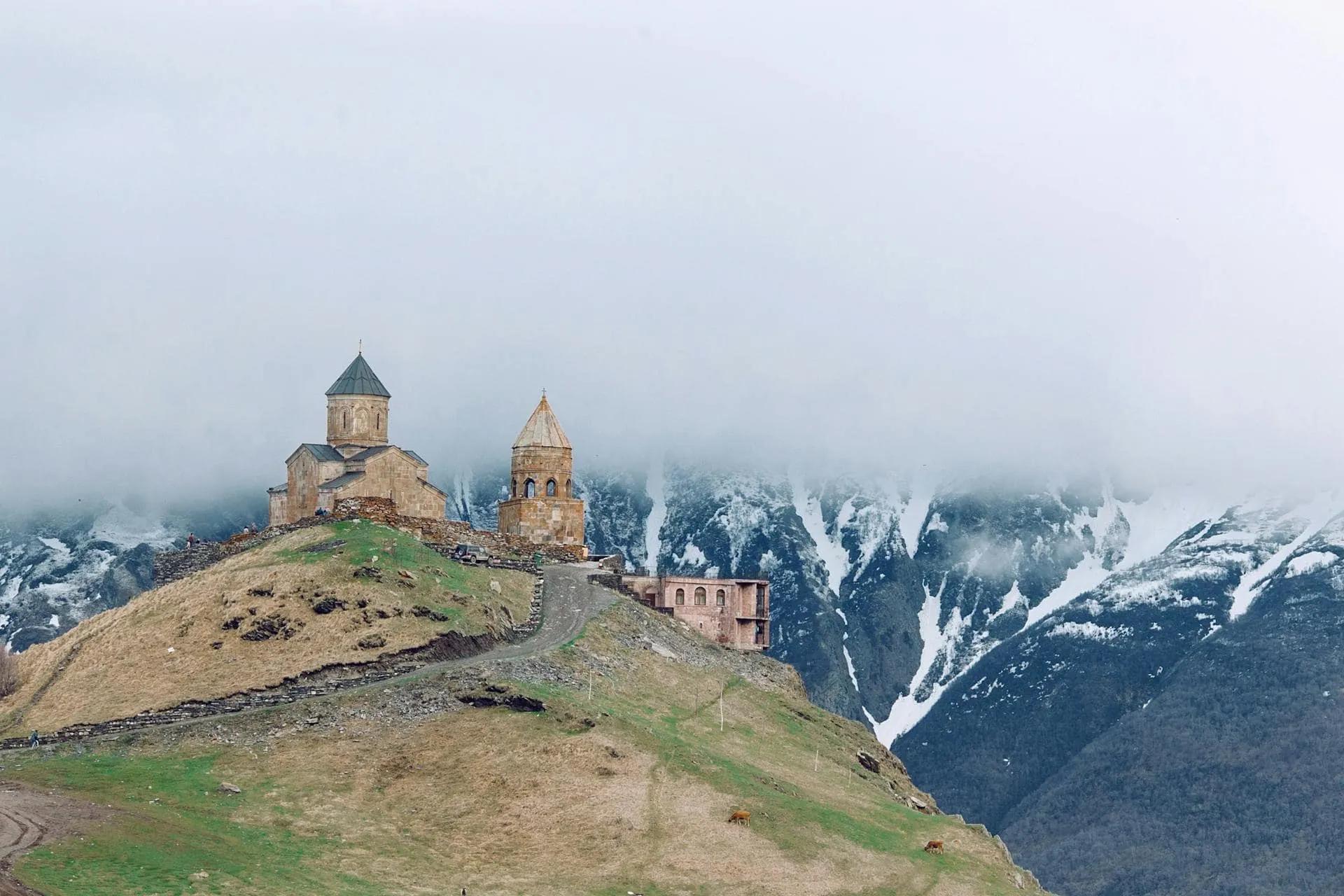 Igreja antiga em montanha nevada, cercada por nuvens e paisagem verde.