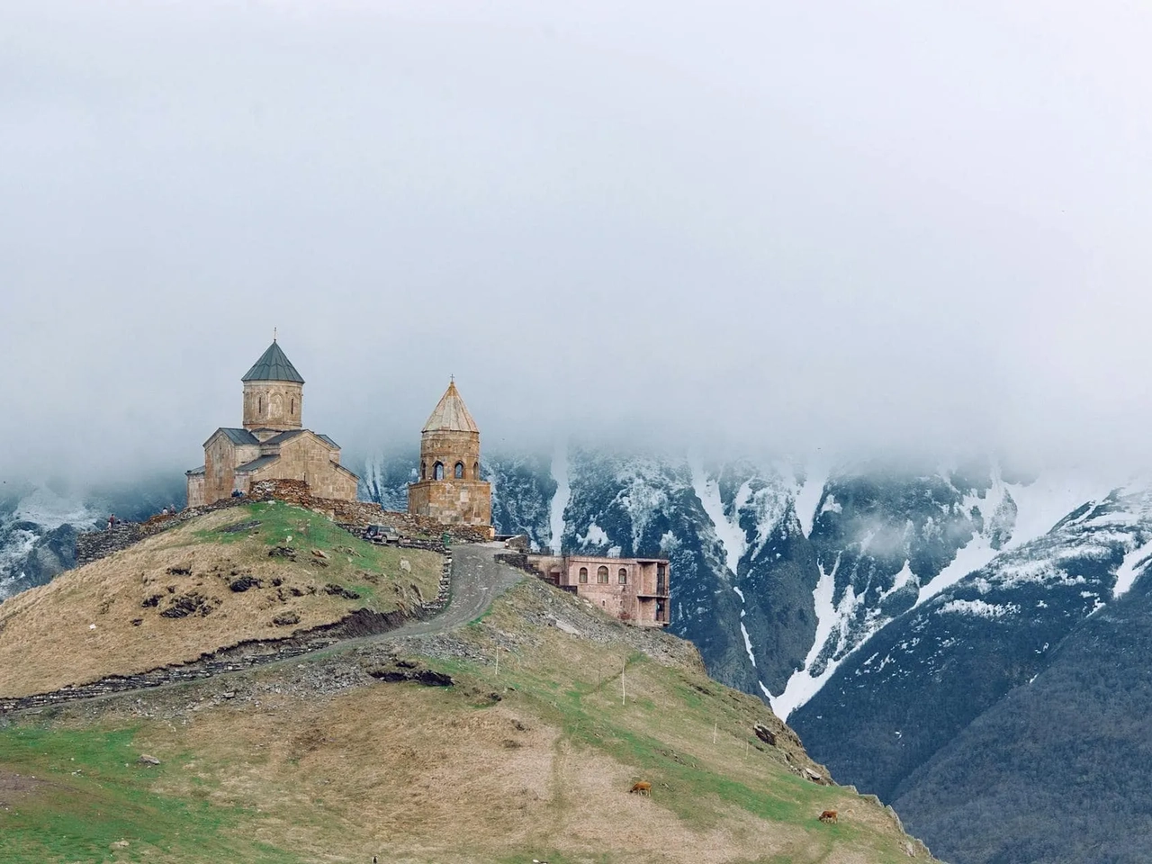Igreja antiga em montanha nevada, cercada por nuvens e paisagem verde.