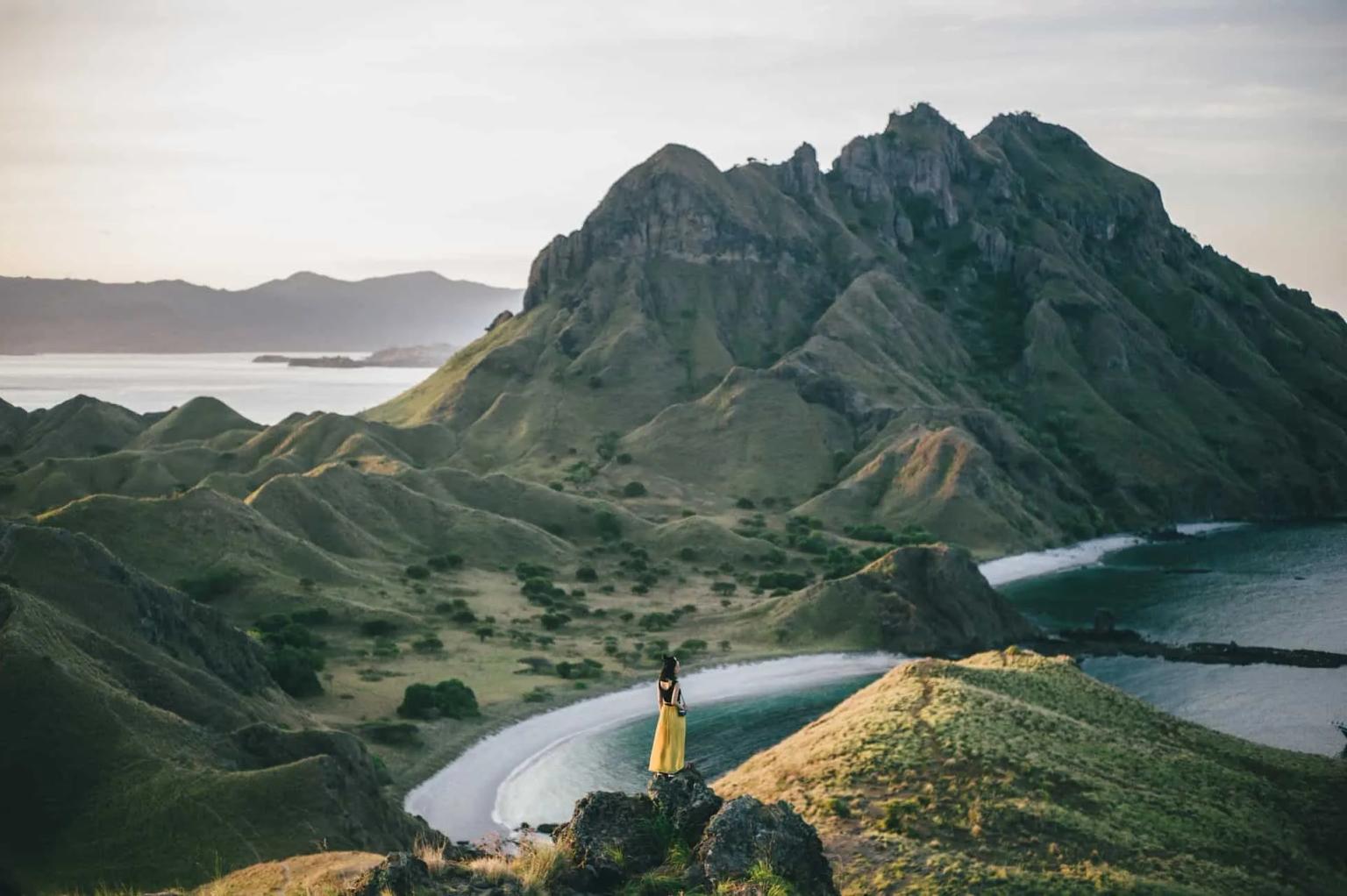 Mulher em vestido amarelo contempla paisagem montanhosa e costa na Indonésia.