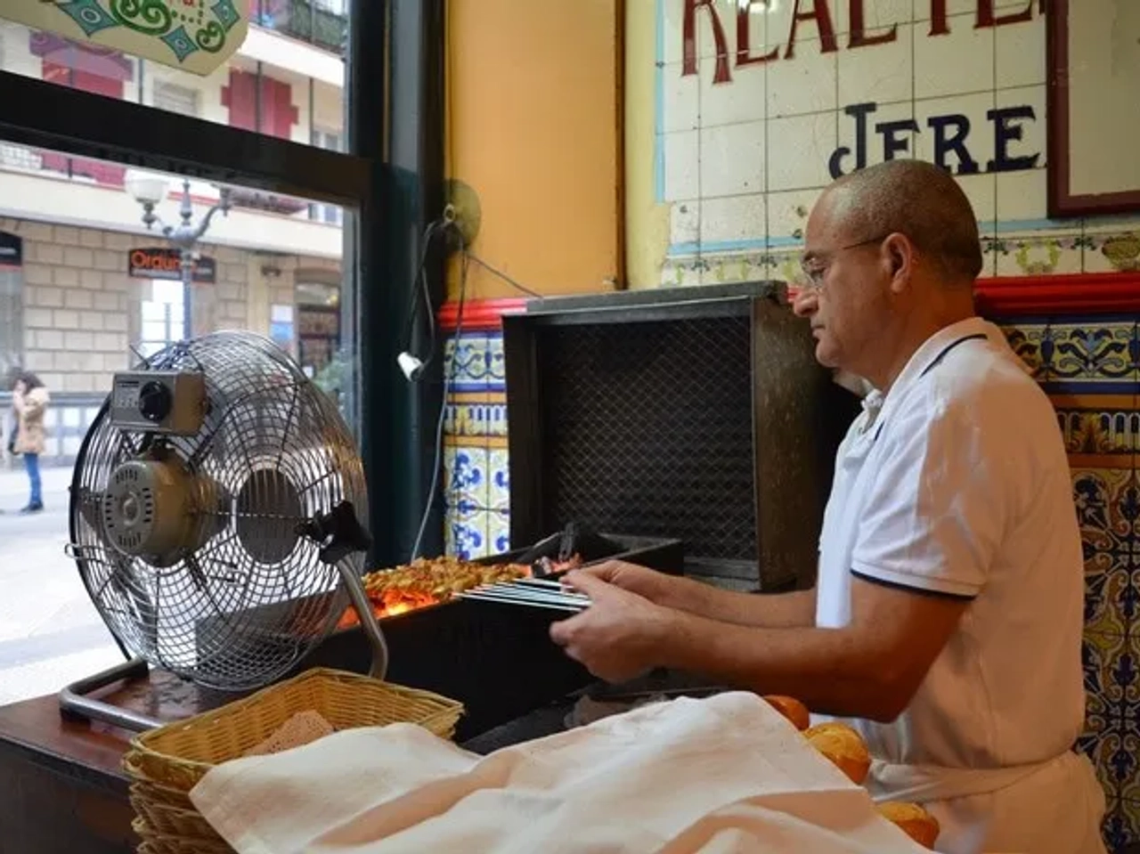 Hombre cocinando pintxos en un bar tradicional de Bilbao. Ventilador y decoración colorida.