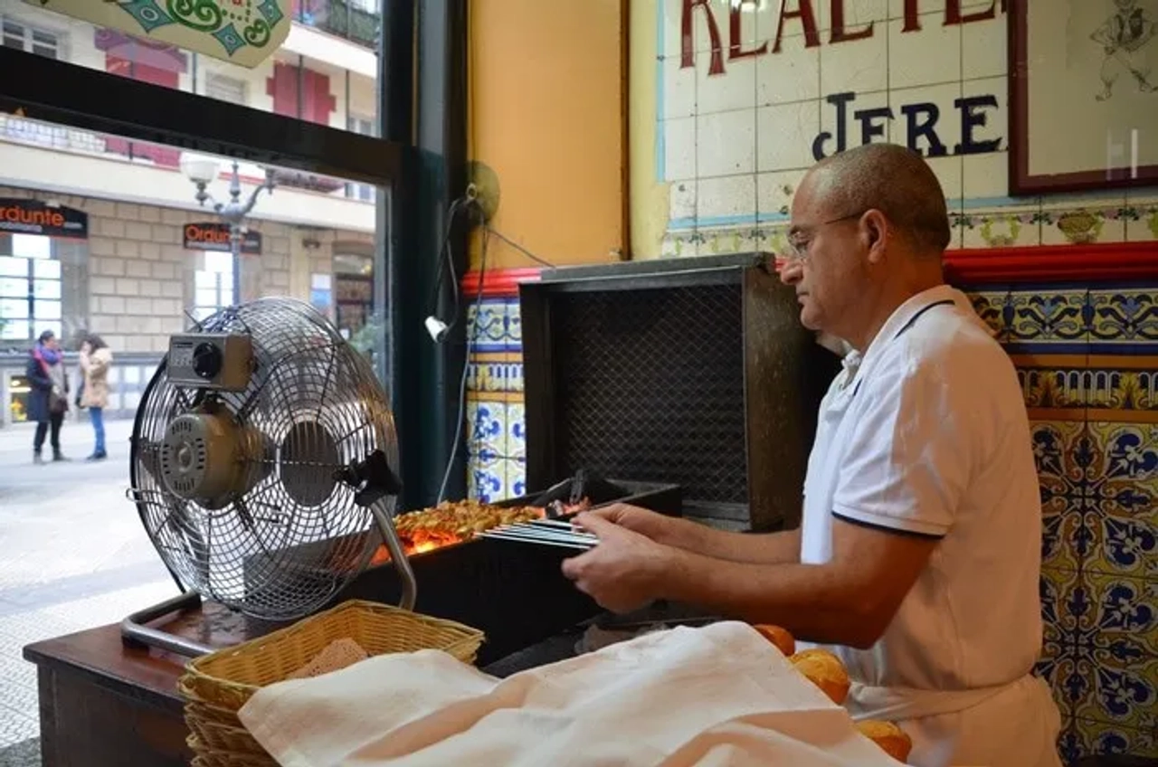 Hombre cocinando pintxos en un bar tradicional de Bilbao. Ventilador y decoración colorida.