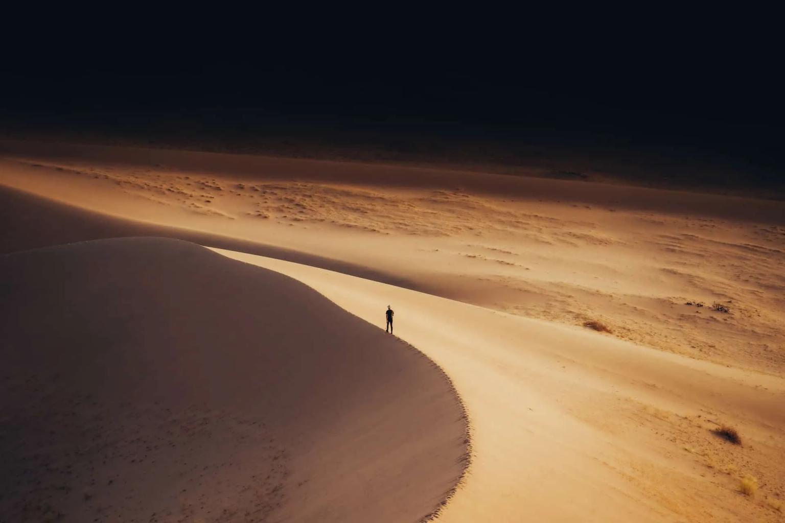 A lone figure stands on a winding sand dune in a vast desert landscape.