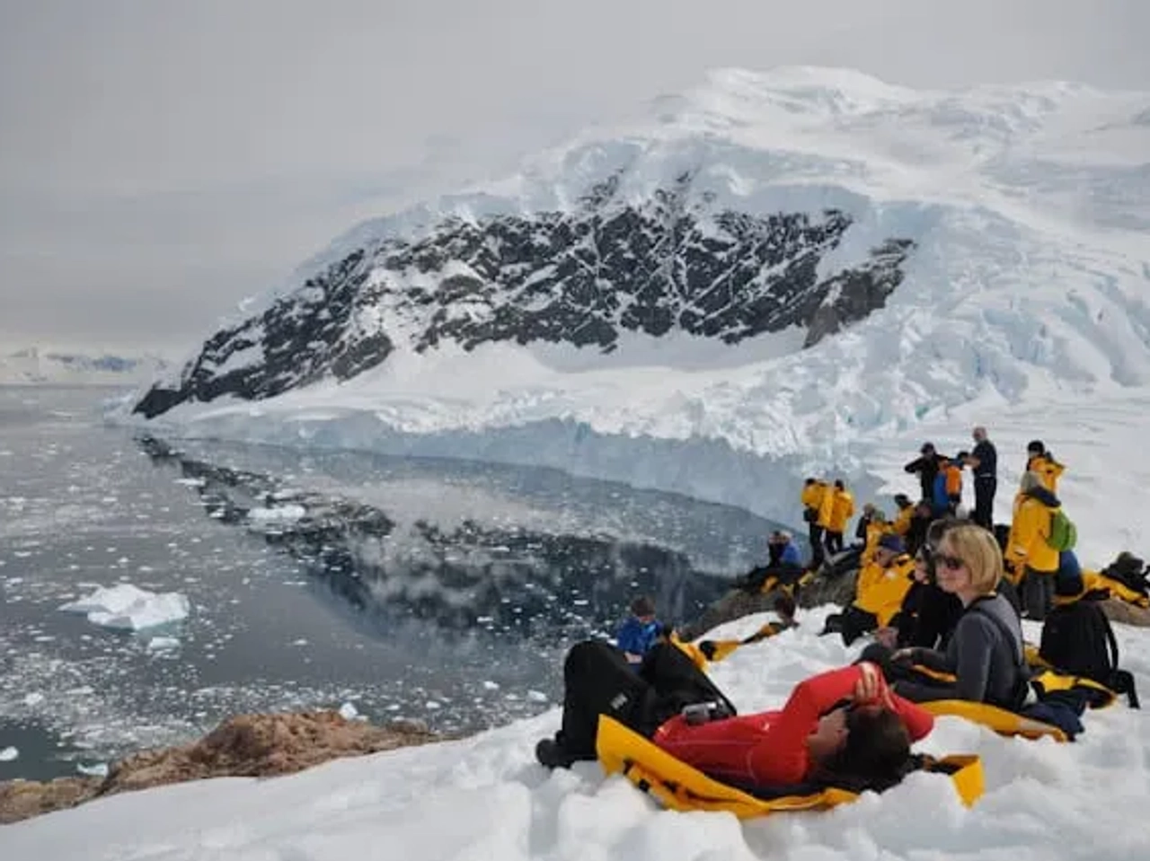 Pessoas relaxando em um cenário gelado da Antártida, com montanhas e água.
