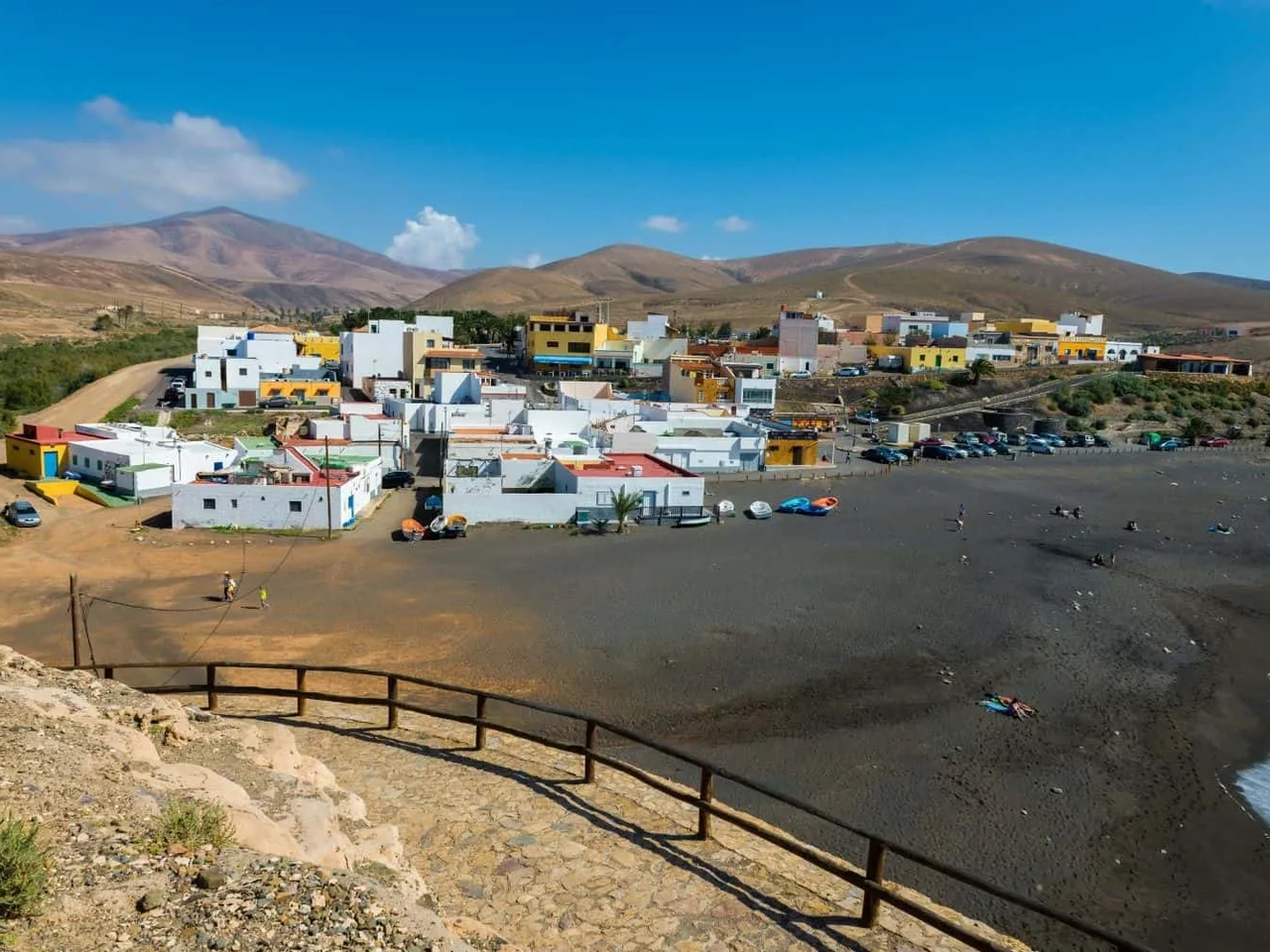 Vista de uma vila costeira nas Ilhas Canárias, com praia e montanhas ao fundo.