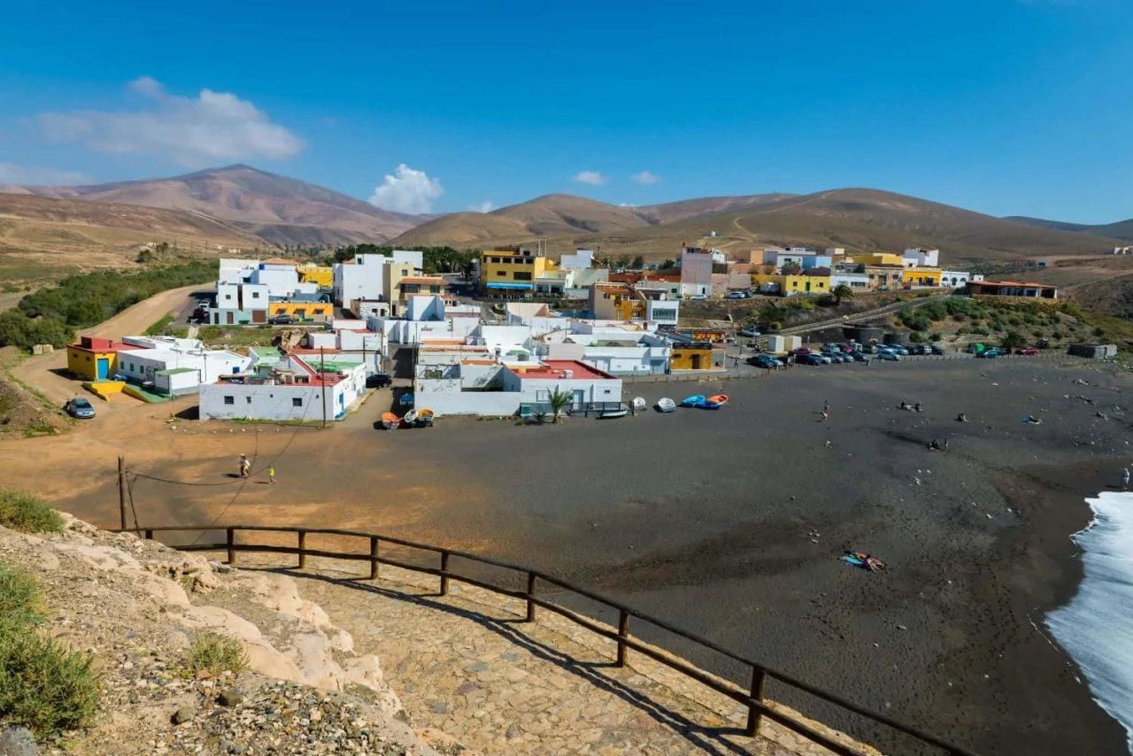 Vista de uma vila costeira nas Ilhas Canárias, com praia e montanhas ao fundo.