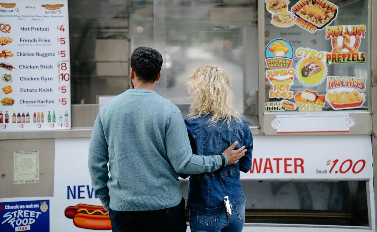 Vista posteriore di una coppia che sceglie degli snack presso un food truck urbano, con varie opzioni di fast food.