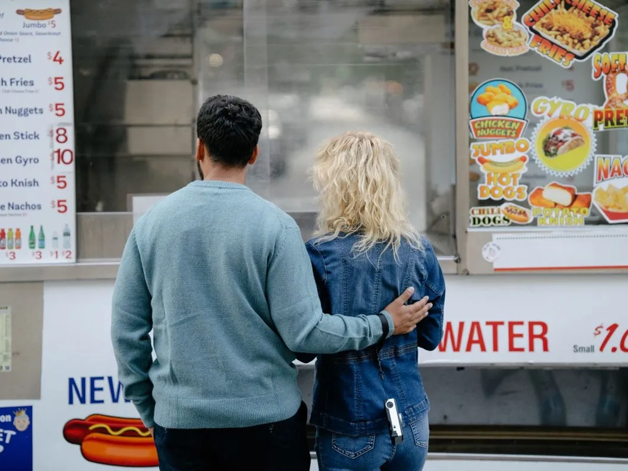 Vista posteriore di una coppia che sceglie degli snack presso un food truck urbano, con varie opzioni di fast food.