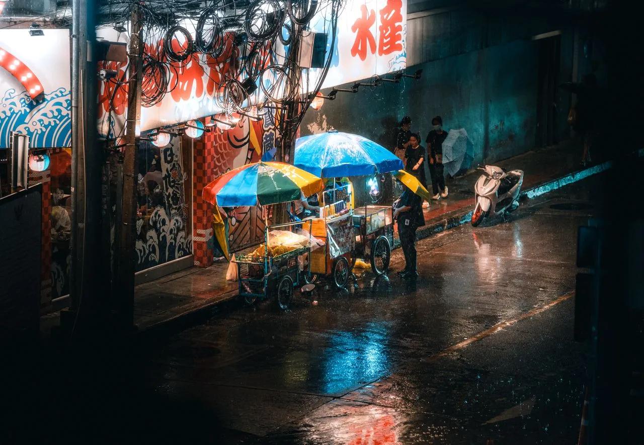 Mercato notturno di Bangkok sotto la pioggia, con bancarelle e ombrelli lungo una strada bagnata.