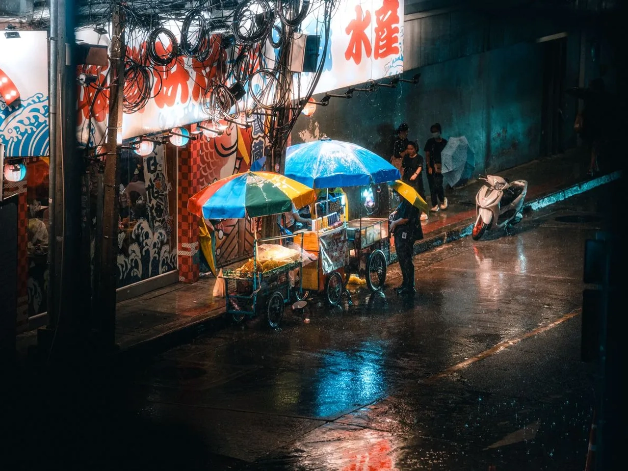 Mercato notturno di Bangkok sotto la pioggia, con bancarelle e ombrelli lungo una strada bagnata.