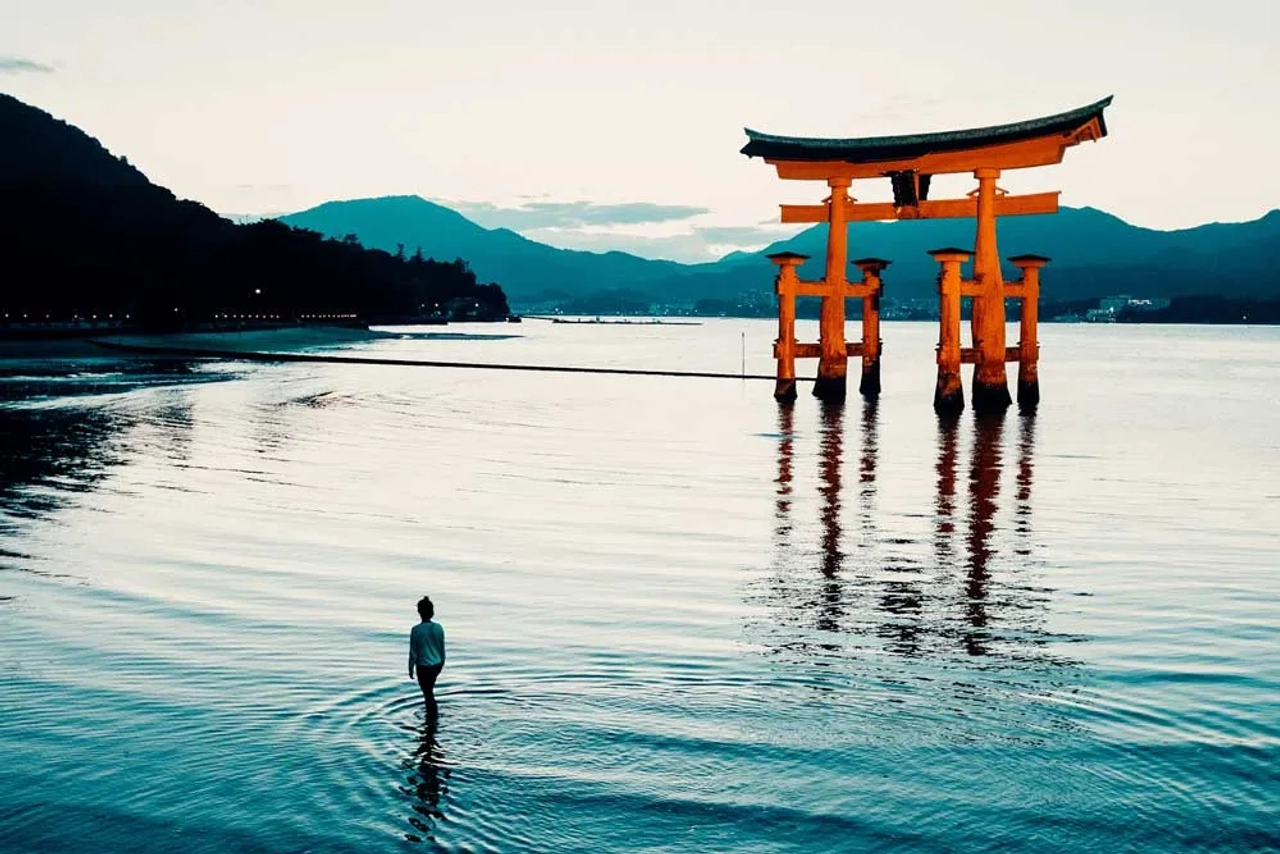 Grande torii rosso del santuario di Itsukushima a Miyajima, riflesso sull’acqua al tramonto, con una persona in piedi sulla riva e le montagne sullo sfondo.