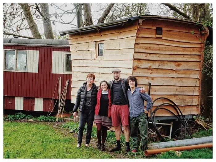 Cuatro personas posan frente a una casa de madera en un entorno natural.