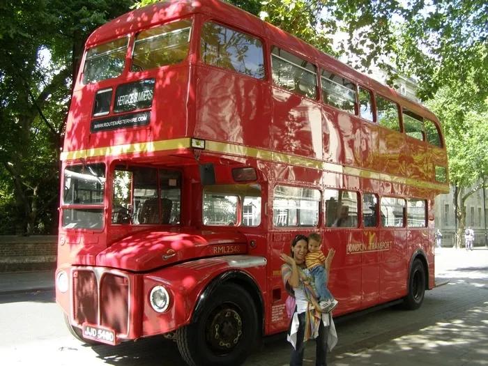 Mulher segurando criança ao lado de um ônibus vermelho de dois andares.