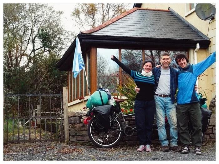 Tres personas sonrientes posan frente a una casa con una bicicleta cargada.