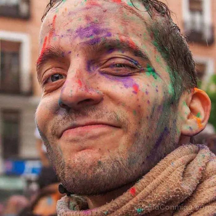 Hombre sonriente con rostro pintado de colores en un ambiente festivo.