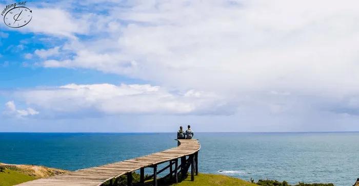 Pareja sentada en un muelle de madera, mirando el mar y el cielo.