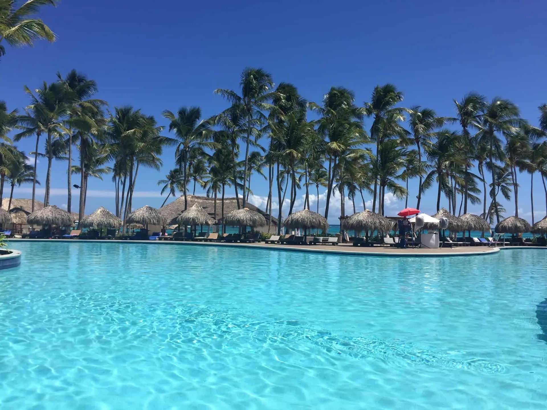 Piscina azul rodeada por palmeiras e cabanas em um resort na República Dominicana.