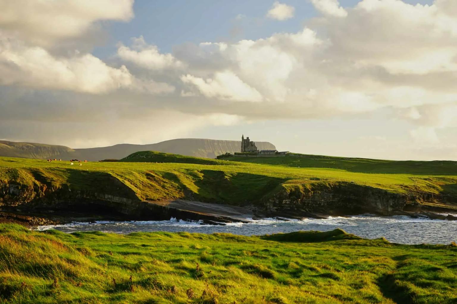Paisagem irlandesa com colinas verdes e um castelo à beira-mar.
