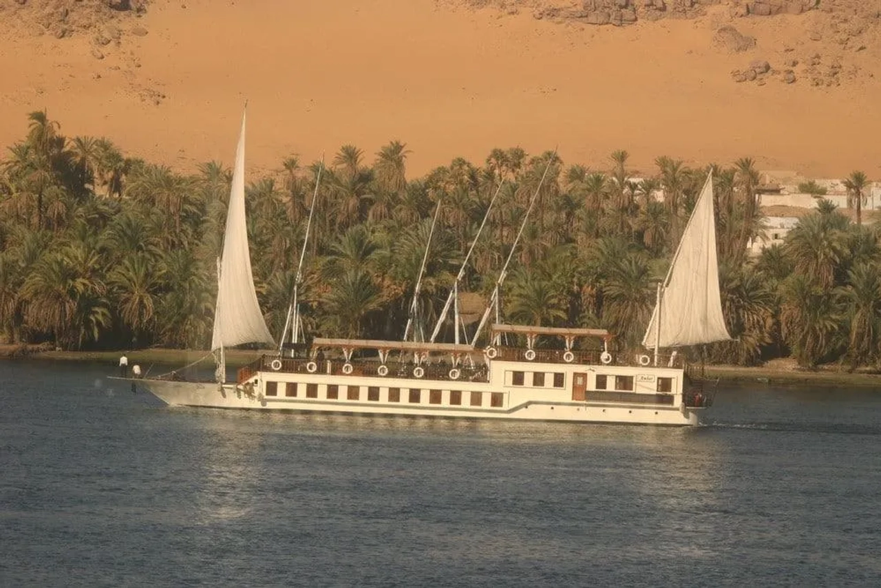 Crociera sul Nilo in Egitto con barca a vela tradizionale, palme lungo la riva e paesaggio desertico sullo sfondo.