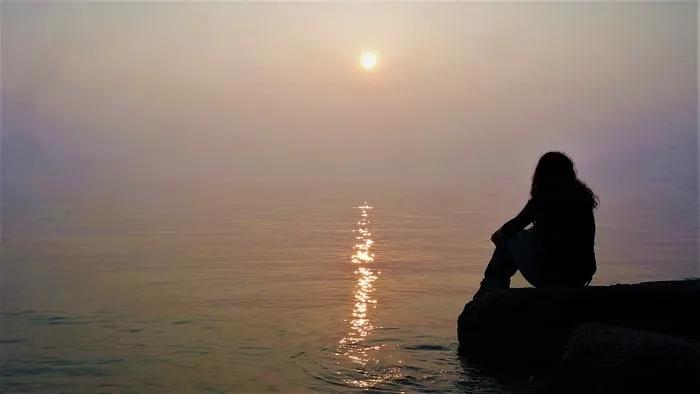 Mujer sentada junto al agua, contemplando un atardecer en un ambiente sereno.