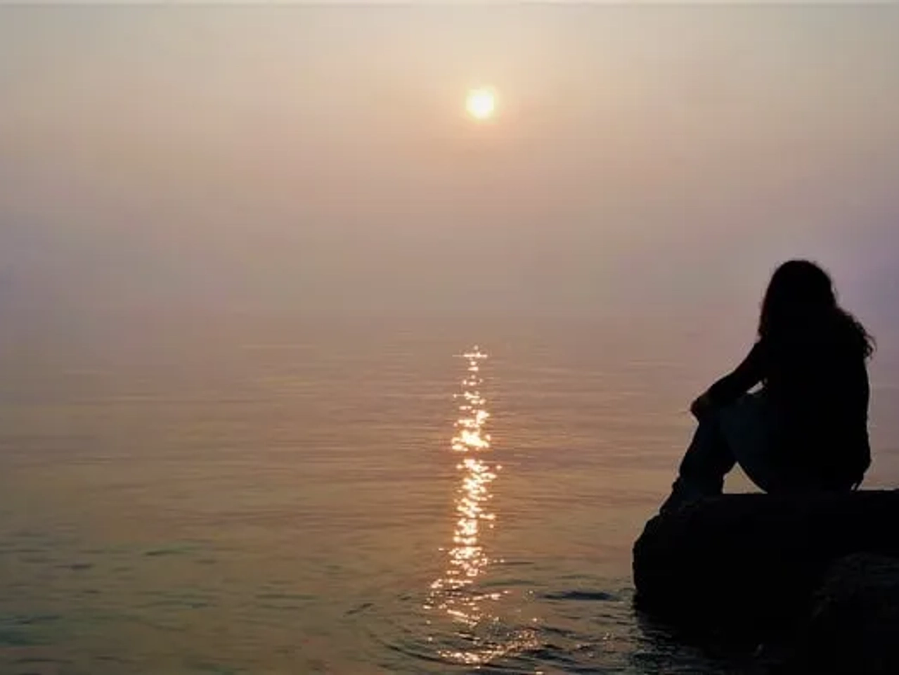 Mujer sentada junto al agua, contemplando un atardecer en un ambiente sereno.