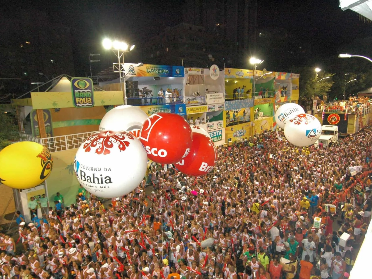 Multidão animada durante o Carnaval, com grandes balões e palcos ao fundo.