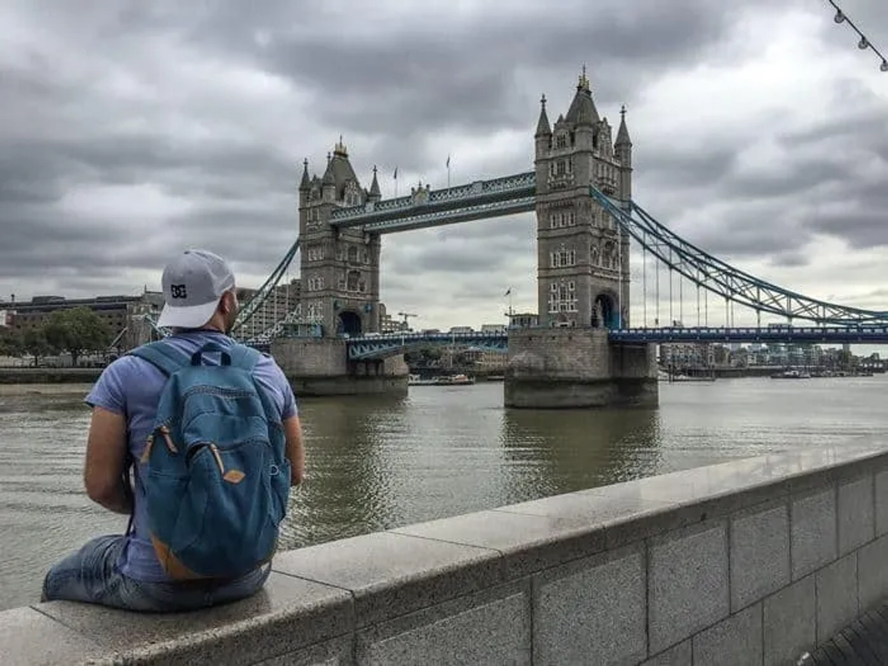 Hombre sentado frente al Puente de la Torre en Londres, con un cielo nublado.