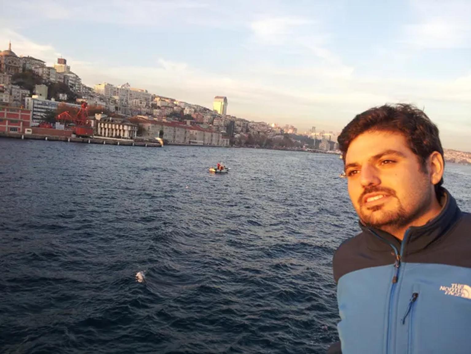 Hombre posando junto al mar, con una ciudad costera al fondo.