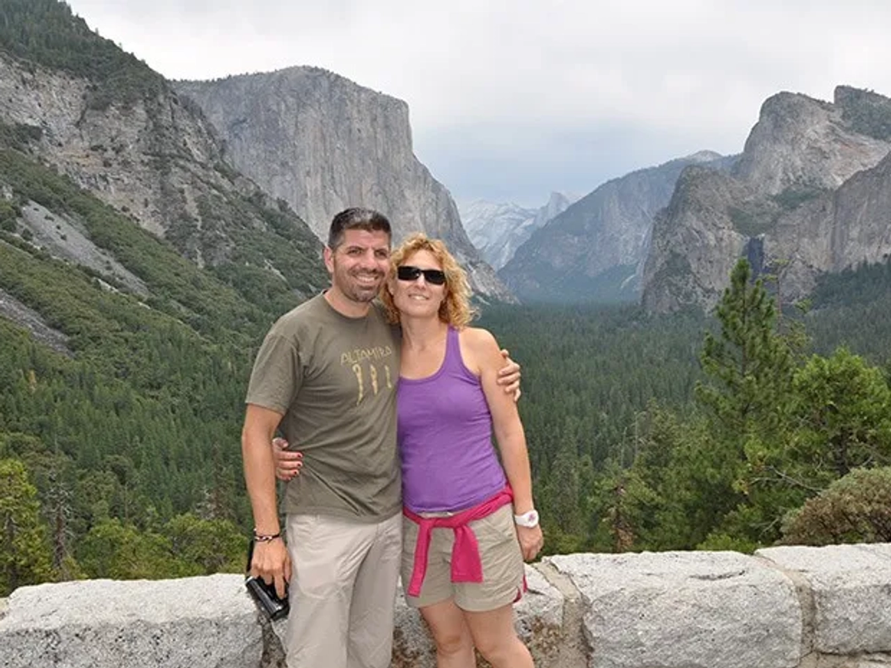 Pareja sonriente en un mirador con montañas y bosque de fondo.