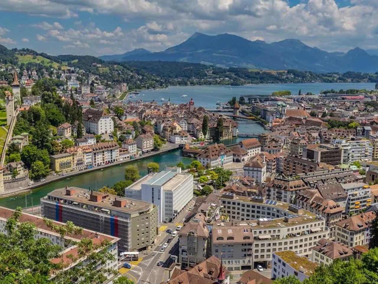 Vista panorâmica de Lucerna, com montanhas e lago ao fundo. Arquitetura charmosa e verde.