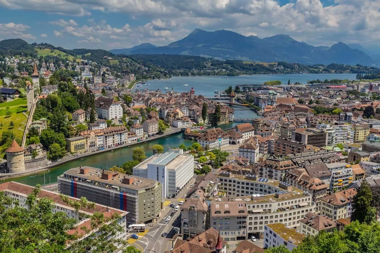 Vista panorâmica de Lucerna, com montanhas e lago ao fundo. Arquitetura charmosa e verde.