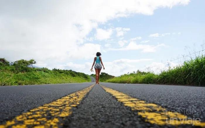 Mujer caminando por una carretera rodeada de naturaleza y cielo despejado.