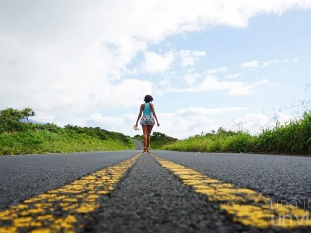 Mujer caminando por una carretera rodeada de naturaleza y cielo despejado.