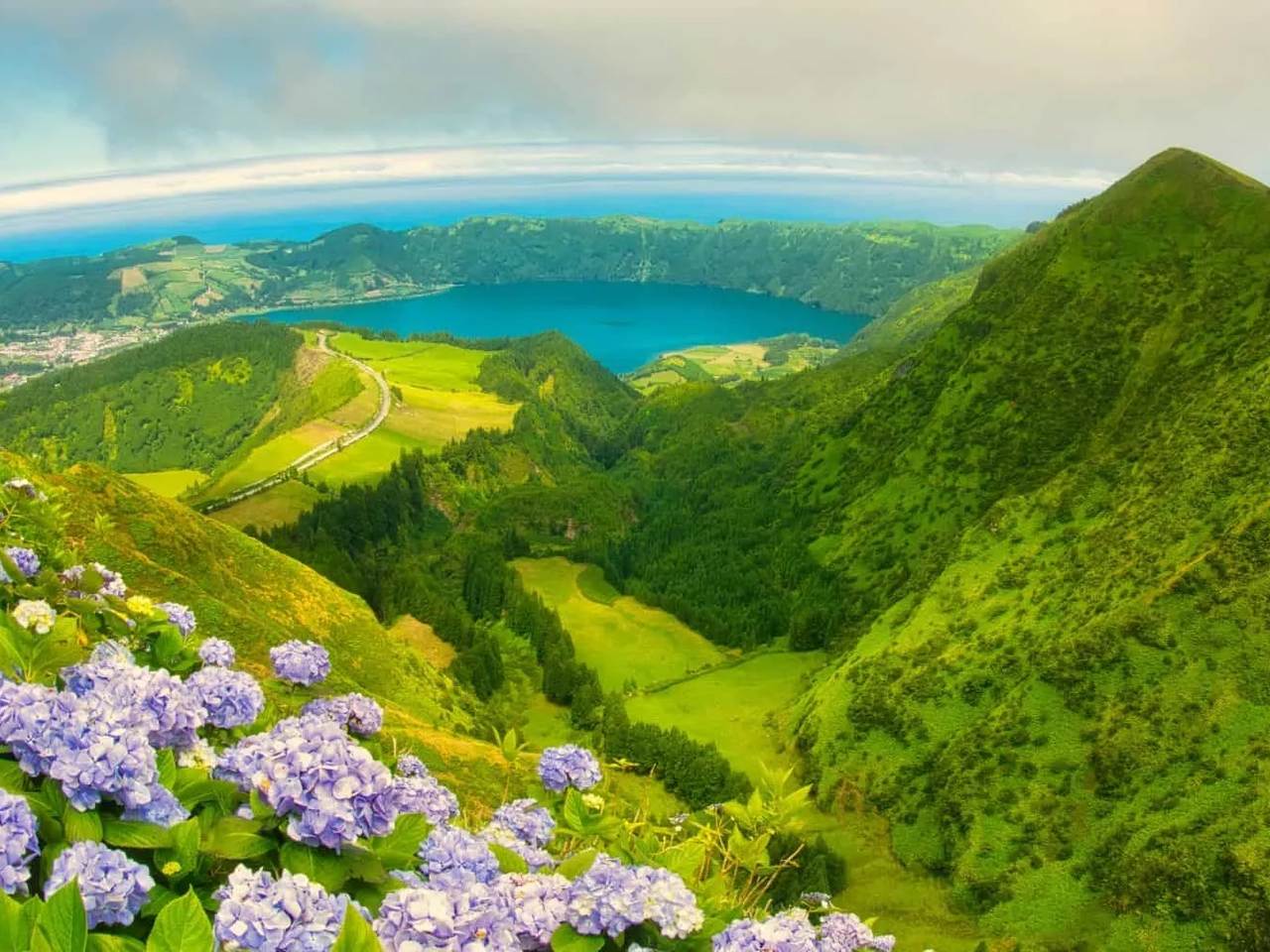 Vista panorâmica de São Miguel, com montanhas verdes e flores roxas em primeiro plano.
